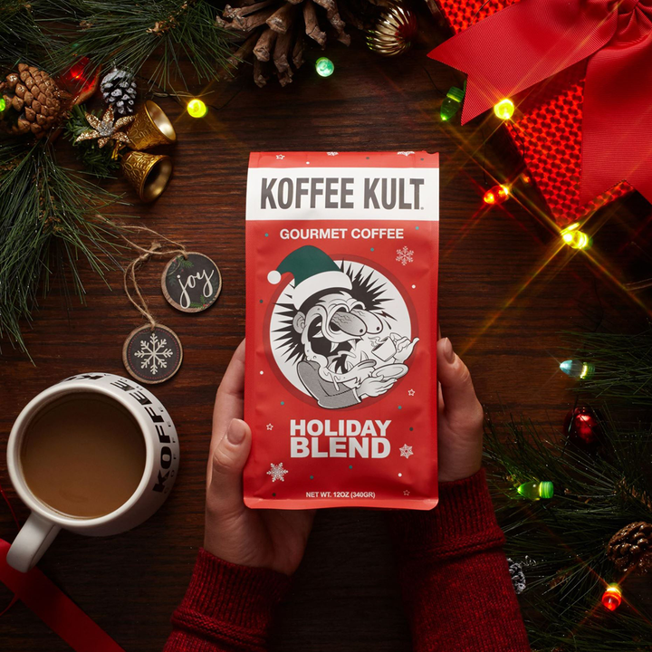 Koffee Kult Holiday Blend coffee package held by a person with Christmas decorations on a wooden surface.