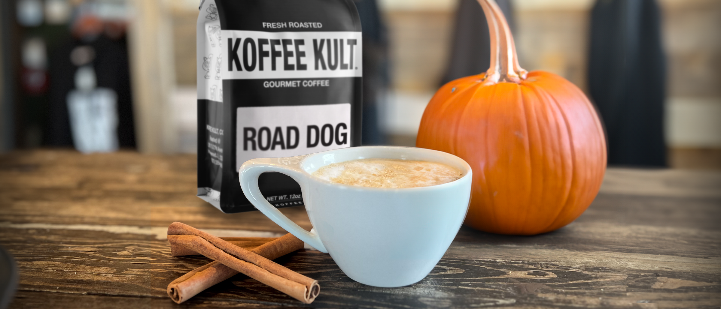 Koffee Kult Road Dog coffee bag with a cup of coffee, pumpkin, and cinnamon sticks on a wooden surface.