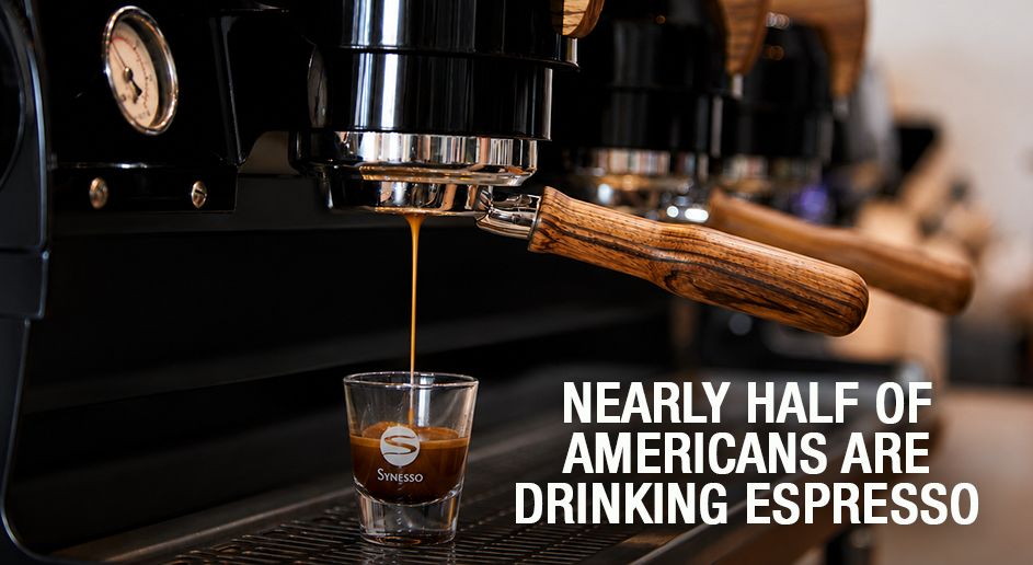 A close-up of a black espresso machine dispensing a stream of espresso into a small clear glass cup with the Smigasso logo. The background is softly blurred, and bold white text on the right reads, 'NEARLY HALF OF AMERICANS ARE DRINKING ESPRESSO.