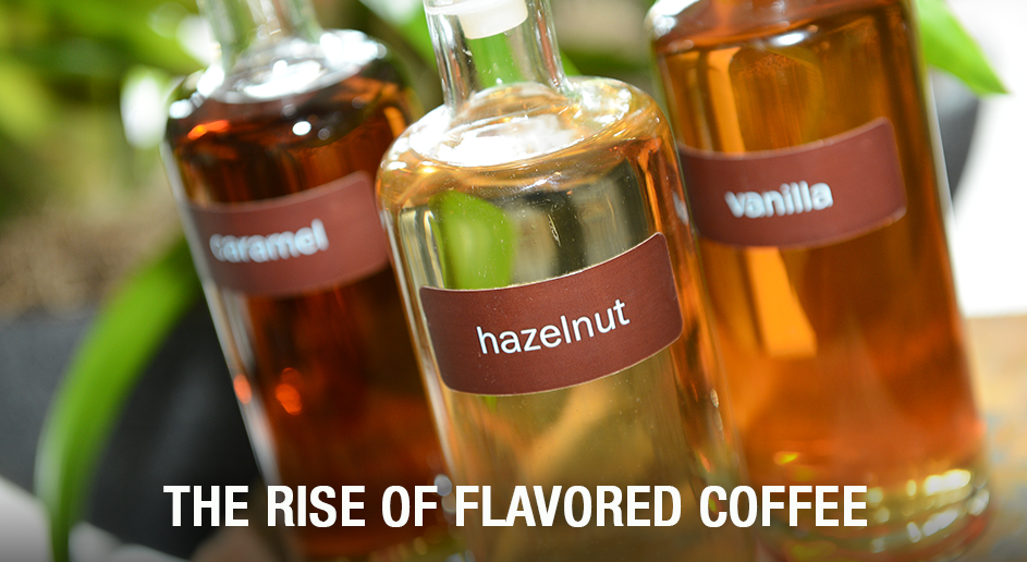 Three clear glass bottles of flavored coffee syrup with labels reading caramel, hazelnut, and vanilla, filled with amber-colored liquid, with a text overlay at the bottom reading 'THE RISE OF FLAVORED COFFEE'.