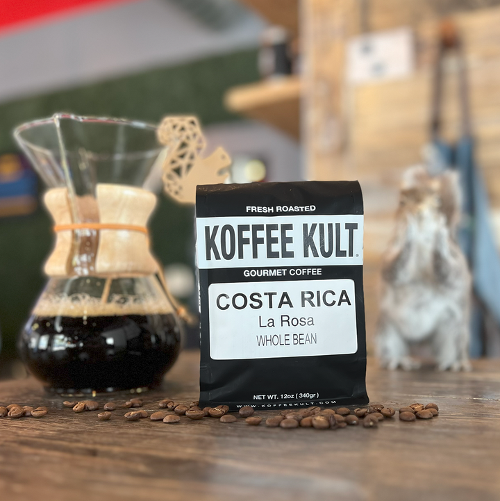 Koffee Kult Costa Rica La Rosa coffee package on a wooden surface with coffee beans and a coffee maker in the background.
