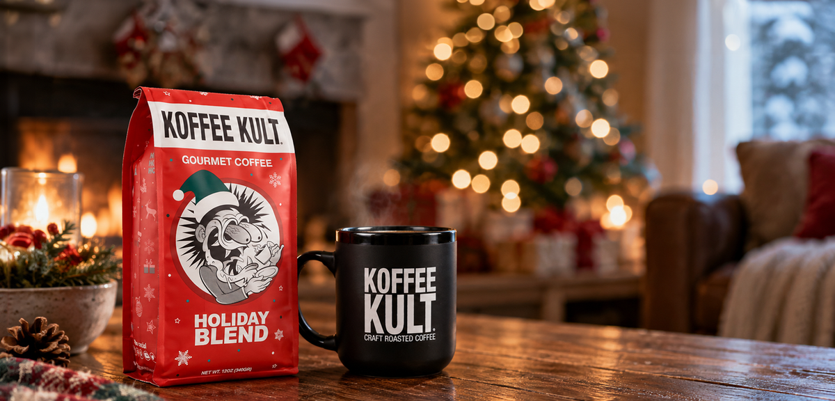 A red bag of Koffee Kult Holiday Blend gourmet coffee and a black ceramic mug with the Koffee Kult logo, set on a wooden table in a cozy, warm holiday living room with a blurred Christmas tree in the background.