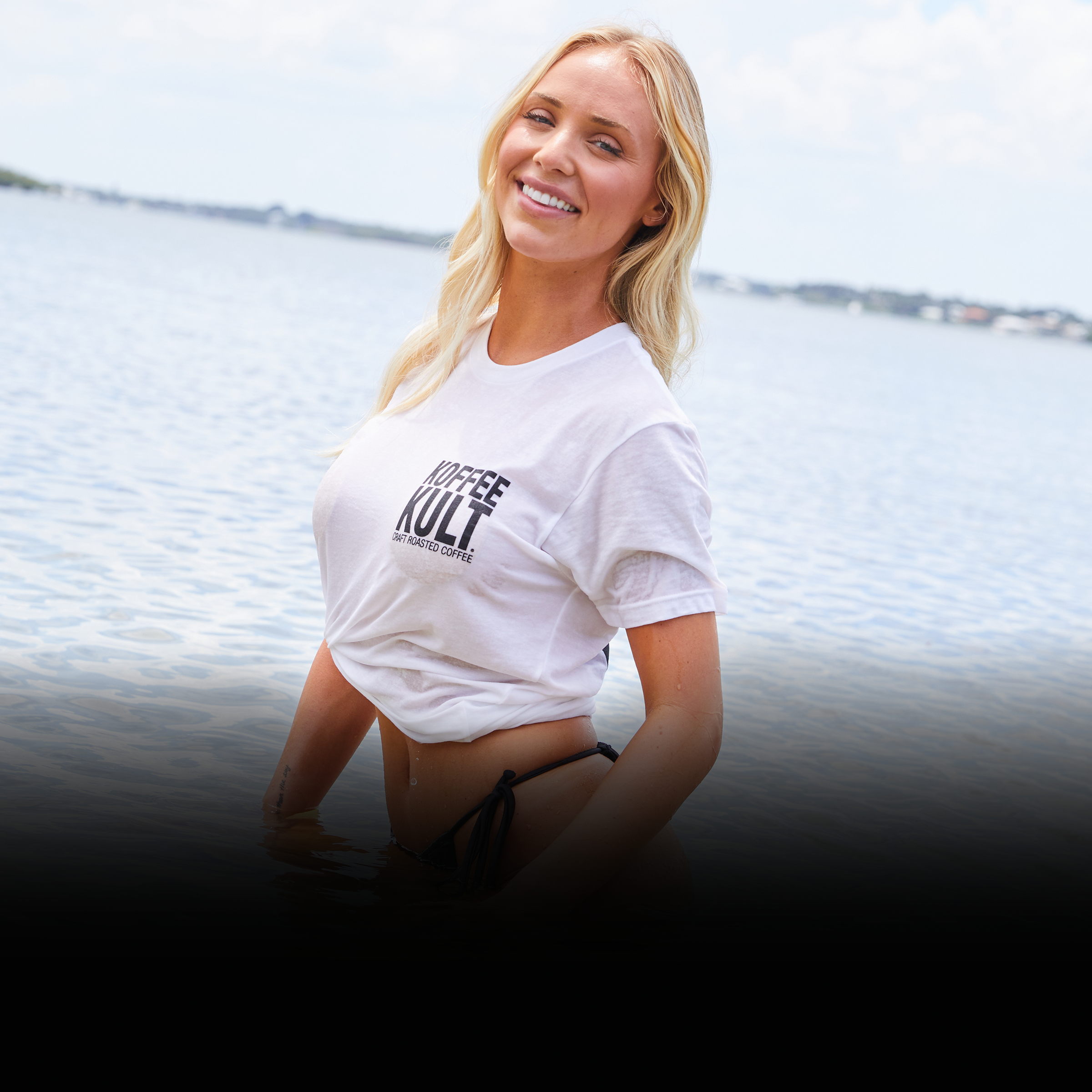 Woman wearing a white t-shirt with 'Koffee Kult' logo by a body of water.