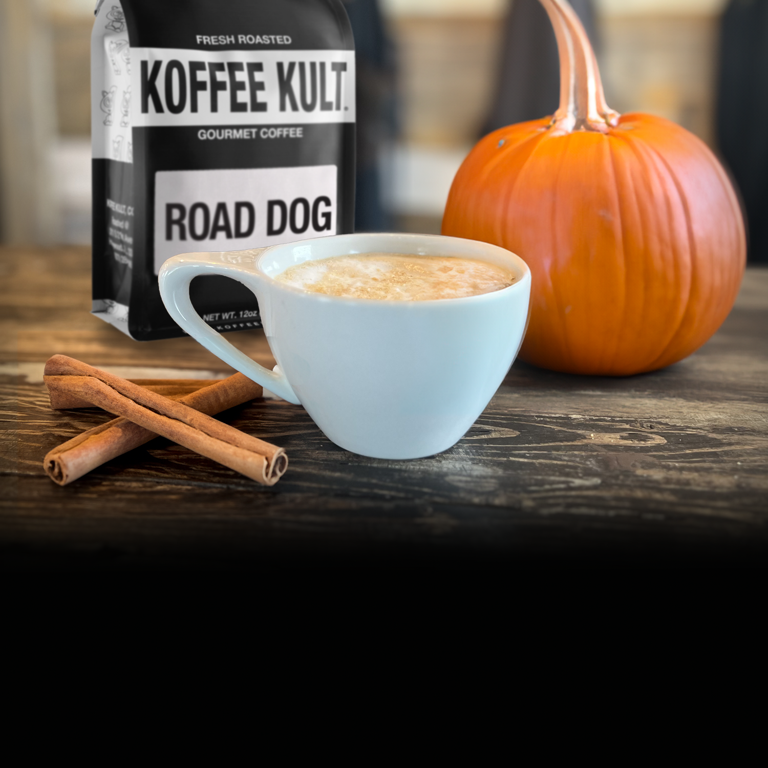 Koffee Kult Road Dog coffee package with a cup of coffee, cinnamon sticks, and a pumpkin on a wooden surface.