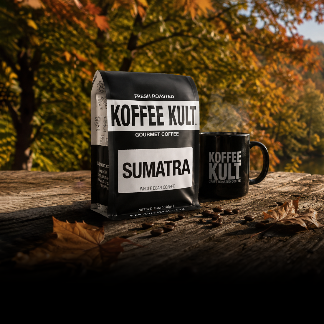 A bag of Koffee Kult Sumatra whole bean coffee stands on a rustic wooden surface next to a matching black coffee mug, with scattered coffee beans and autumn leaves, set against a blurred background of colorful fall foliage.